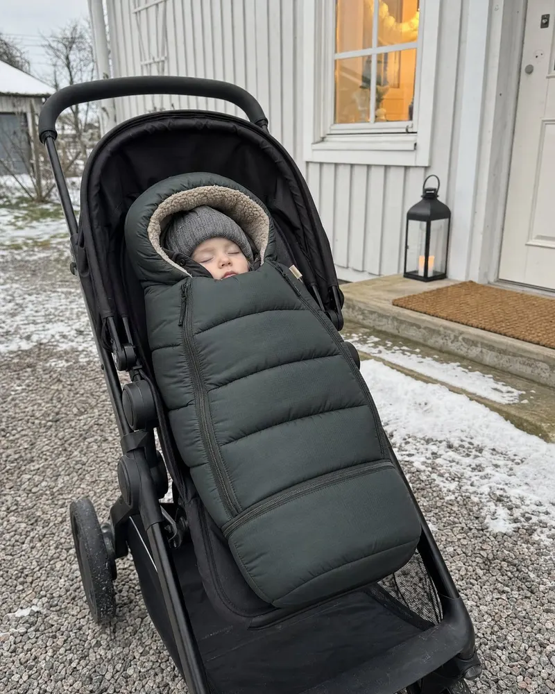 Sovande barn i en mörkgrön åkpåse i en barnvagn utanför ett hus en vinterdag